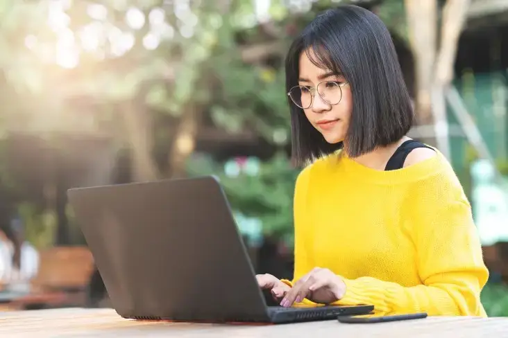 Young woman with short bob haircut wearing yellow sweater and glasses typing on laptop outdoors