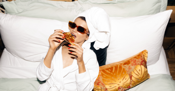 Celebrity Woman wears sunglasses and robe in bed. Her hair is wrapped in towel after esthetician spa treatment. She eats pizza