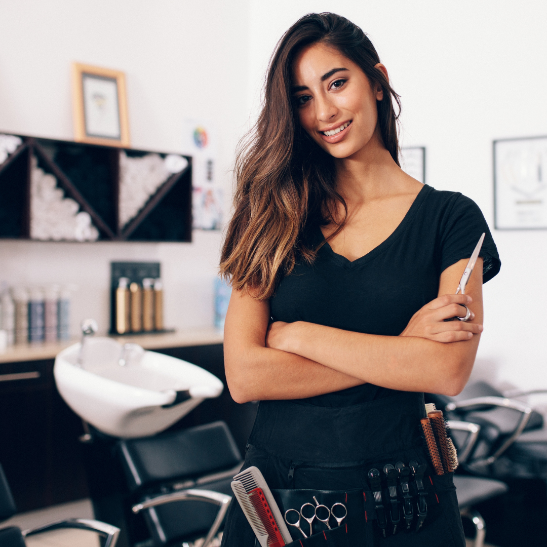 Cosmetology school student with arms crossed smiles wearing tool belt